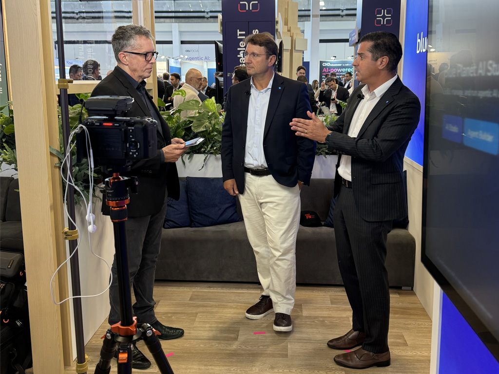 mosaic_DTW4 Three conference attendees participate in an on-camera discussion at the Blue Planet booth, with recording equipment visible in the foreground.