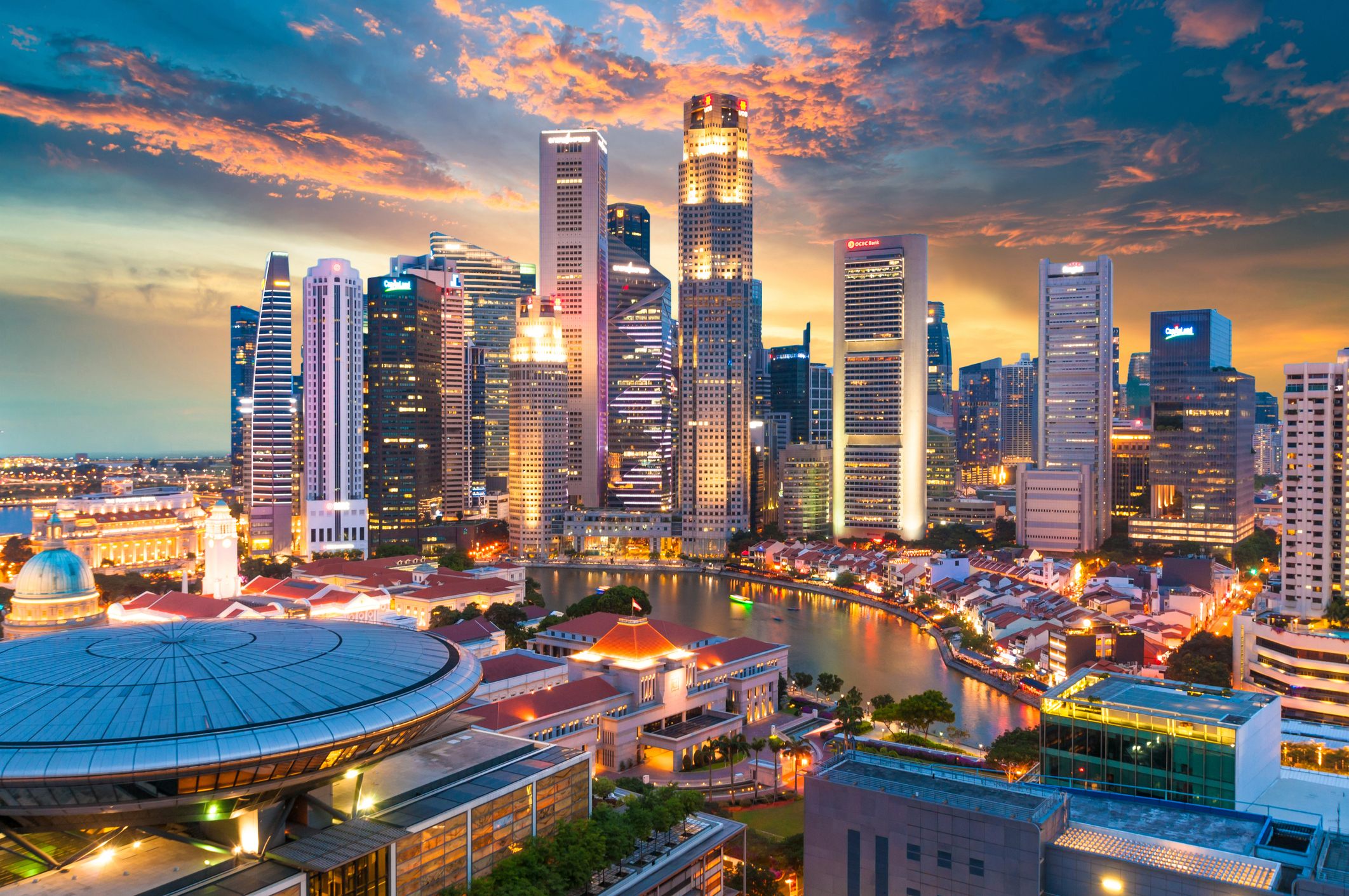 Panoramic view of downtown Singapore at dusk, with high-rise office buildings, waterfront canals, and city lights glowing beneath a vibrant sunset sky.