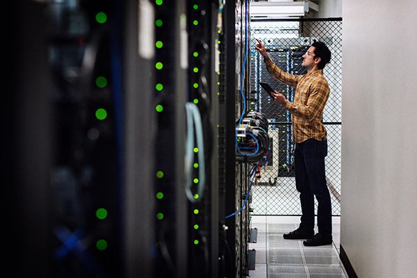 Engineer inspecting network equipment in a server room, holding a tablet among rows of active racks.
