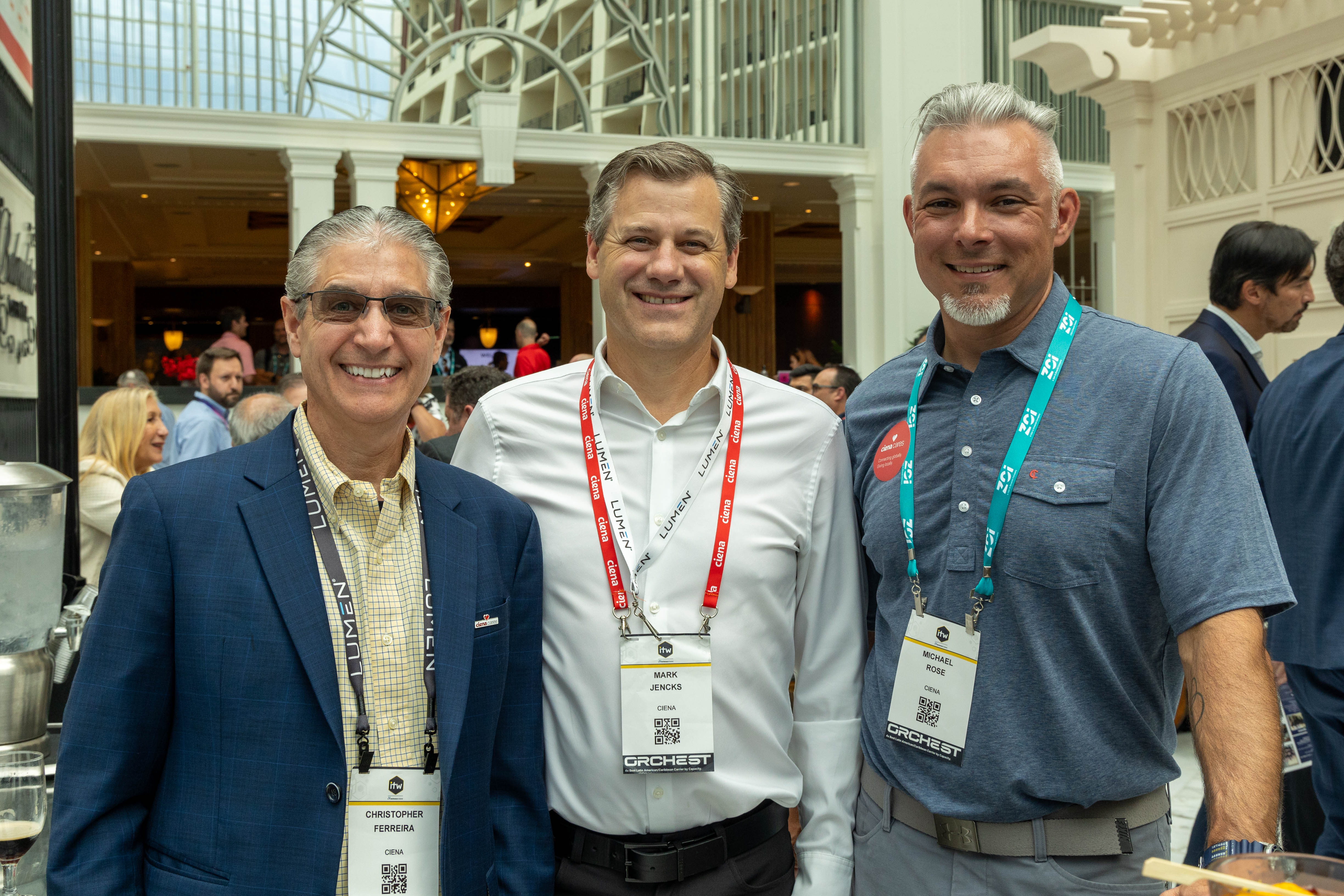 Three event attendees wearing conference badges smile for a photo in a hotel atrium during a networking event associated with the Ciena Cares mobility restoration program.