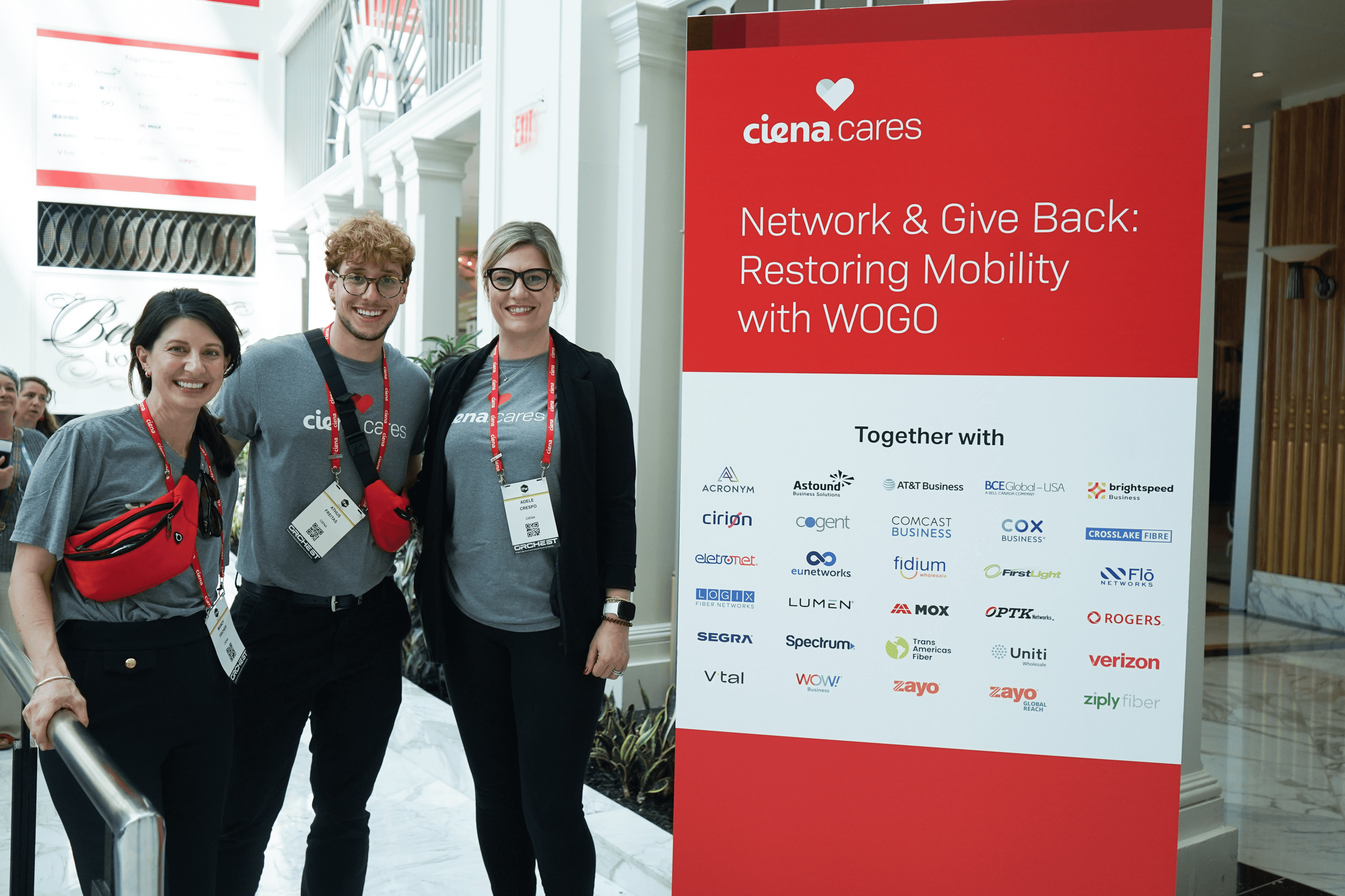Volunteers standing beside a Ciena Cares “Restoring Mobility with WOGO” event sign.