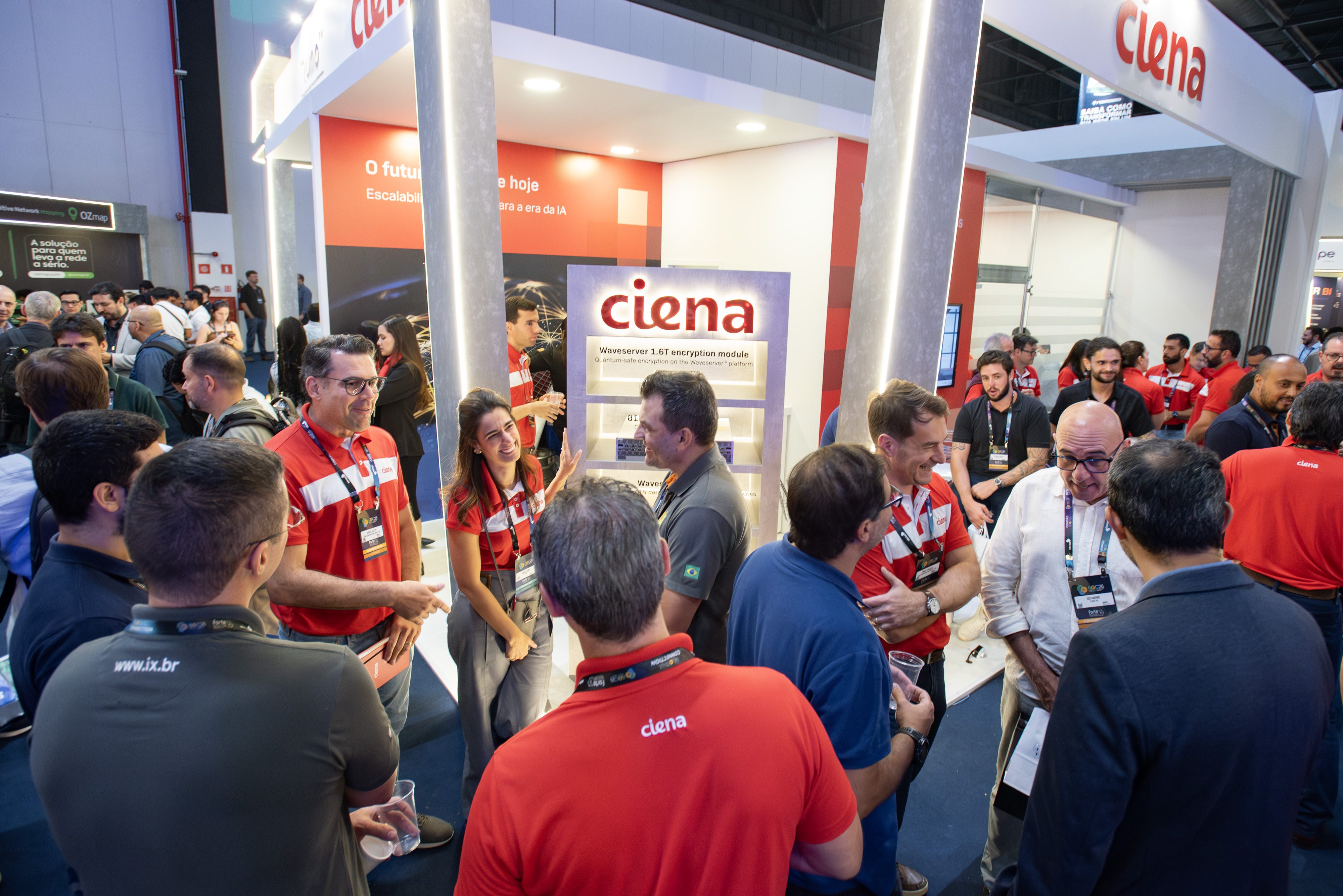 A crowded trade-show booth with many attendees networking and discussing technology. Multiple staff members wearing red-and-white Ciena-branded shirts.
