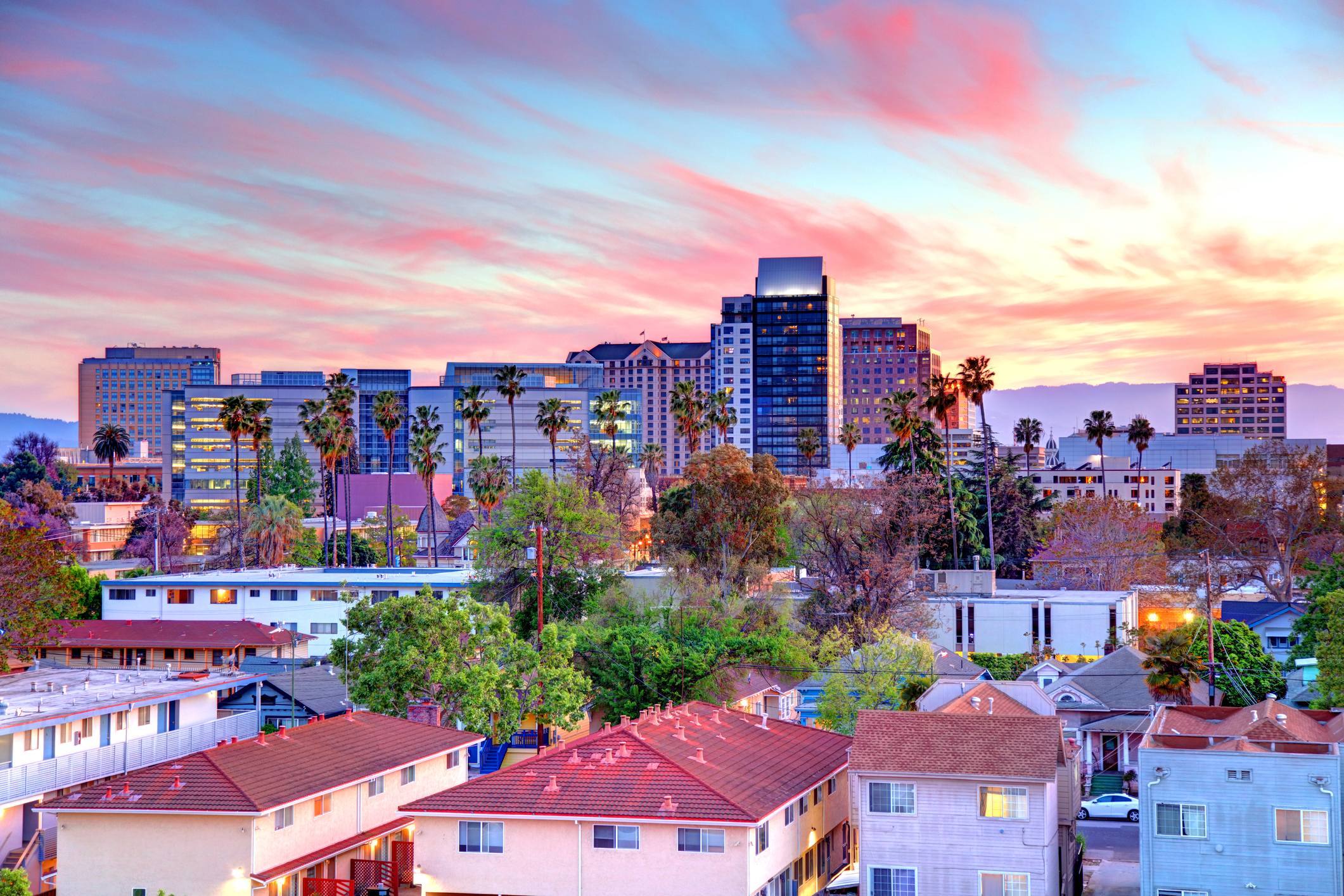 Downtown San Jose skyline with palm trees and residential rooftops under a vibrant sunset sky.
