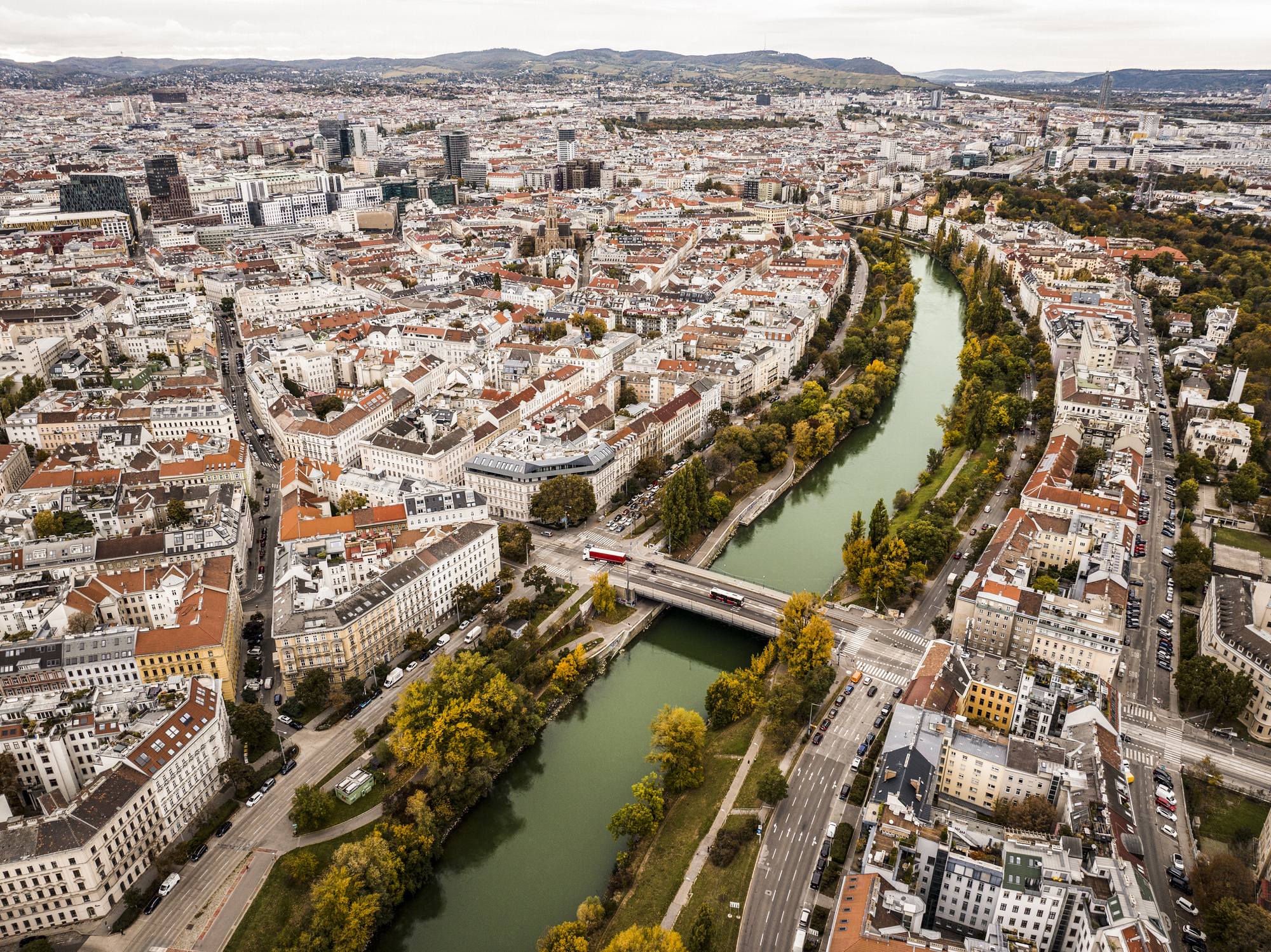 Aerial view of Vienna, Austria, showing the Danube Canal winding through the city, with historic buildings, tree-lined streets, bridges, and dense urban development extending toward surrounding hills.